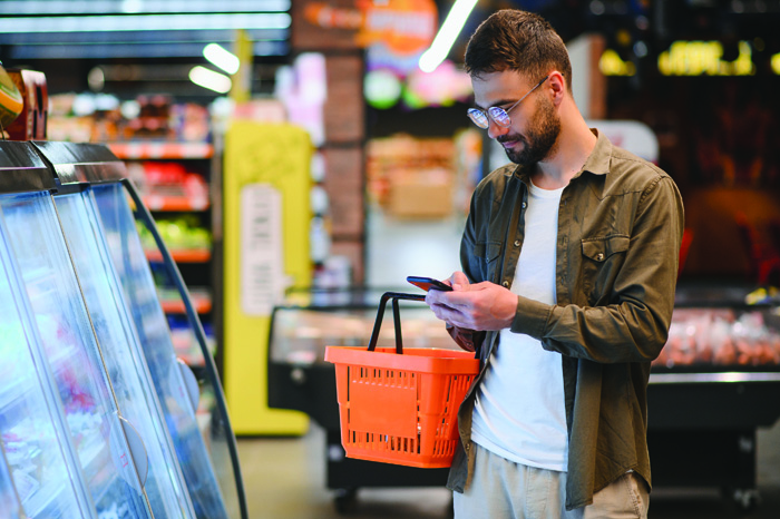 Quick text during shopping. Handsome young man holding mobile phone and smiling while standing in a food store Laut den Prognosen des HDE behalten die Verbraucher erst einmal eine gewisse Konsumzurückhaltung bei. Bild: Adobe Stock